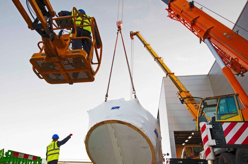 Workers in high-vis directing a crane carrying a heavy machine part.